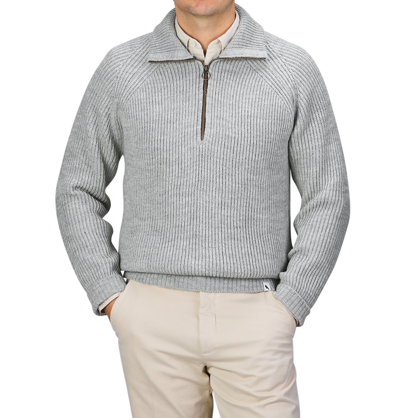 A person stands against a neutral backdrop, hands in pockets, donning a sleek light grey wool zip neck sweater by Peregrine and paired with beige pants.