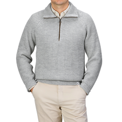 A person stands against a neutral backdrop, hands in pockets, donning a sleek light grey wool zip neck sweater by Peregrine and paired with beige pants.