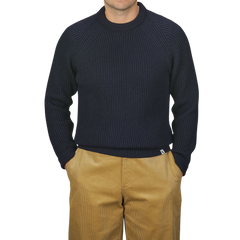 A person wearing a Peregrine Navy Blue Wool Ford Raglan Crewneck sweater and tan pants stands against a neutral background with their hands in their pockets.