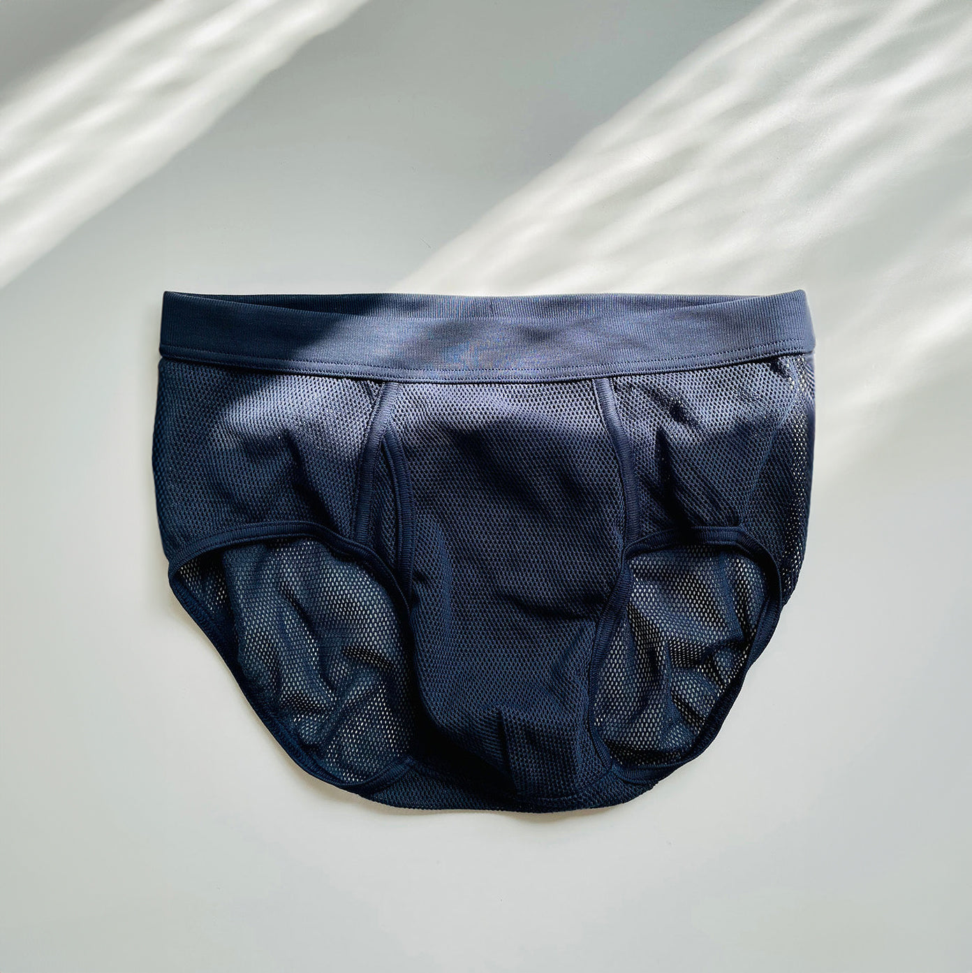 A pair of Platan Organic Cotton Mesh Briefs by The White Briefs, in dark blue and regular fit, rests on a light surface with diagonal sunlight casting gentle shadows, highlighting the classic elegance of organic Pima cotton.