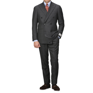 A person sporting the Ring Jacket Grey Calm Twist Wool DB Suit is elegantly poised against a white background. The suit, featuring a full canvas construction and crafted from fine wool fabric, is paired with a crisp white shirt, an orange patterned tie, and classic brown loafers.