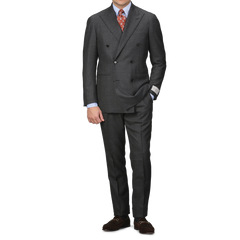 A person sporting the Ring Jacket Grey Calm Twist Wool DB Suit is elegantly poised against a white background. The suit, featuring a full canvas construction and crafted from fine wool fabric, is paired with a crisp white shirt, an orange patterned tie, and classic brown loafers.