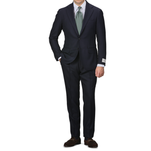 A person in the Navy Blue Calm Twist Wool Suit by Ring Jacket, complemented with a green tie and brown shoes, stands against a plain background, highlighting the elegance of full canvas construction in high-twist wool fabric.