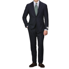 A person in the Navy Blue Calm Twist Wool Suit by Ring Jacket, complemented with a green tie and brown shoes, stands against a plain background, highlighting the elegance of full canvas construction in high-twist wool fabric.