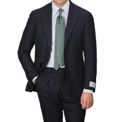 A man is standing against a plain background, wearing the Navy Blue Calm Twist Wool Suit by Ring Jacket, paired with a white shirt and a green tie, one hand casually tucked in his pocket.