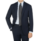 Man in a Ring Jacket Navy Blue High Twist Wool Suit with a light blue shirt and gray tie, standing with one hand in his pocket.