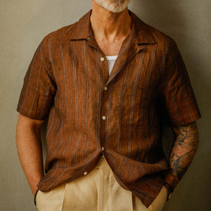 Man in a Universal Works Rust Brown Striped Linen Road Camp Collar Shirt and beige linen pants with visible tattoo on his arm.