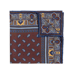 A neatly folded, square-shaped Chocolate Recioto Silk Pocket Square from Sera Fine Silk showcases a vibrant paisley pattern in blue, brown, and yellow tones with one corner visible. This luxury silk pocket square is made in Italy.