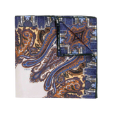 The Cortina Blue Silk Pocket Square by Sera Fine Silk is a folded silk accessory featuring an intricate paisley pattern in blue, orange, and brown colors on a white background. It boasts elegant hand-rolled edges.