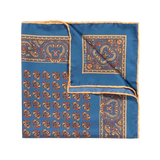 The Nutmeg Montepulciano Silk Pocket Square by Sera Fine Silk is a folded blue handkerchief, crafted from exquisite Como silk, showcasing an intricate orange and gold paisley and geometric pattern against a white background. This seasonal pocket square also features hand-rolled edges for a refined finish.