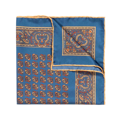 The Nutmeg Montepulciano Silk Pocket Square by Sera Fine Silk is a folded blue handkerchief, crafted from exquisite Como silk, showcasing an intricate orange and gold paisley and geometric pattern against a white background. This seasonal pocket square also features hand-rolled edges for a refined finish.