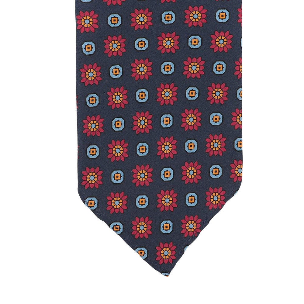 A detailed view of the Stefano Cau Dark Blue Flower Printed Silk Unlined Tie, made from luxurious Italian Como silk, is set against a blue background and features a pattern of red and orange flowers within squares.