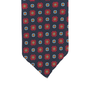 A detailed view of the Stefano Cau Dark Blue Flower Printed Silk Unlined Tie, made from luxurious Italian Como silk, is set against a blue background and features a pattern of red and orange flowers within squares.