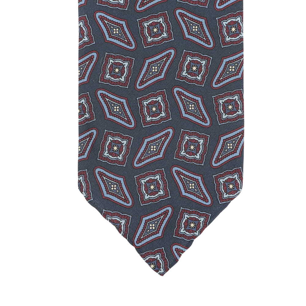 A close-up of a Stefano Cau necktie crafted from luxurious Italian silk, featuring a geometric pattern with red, blue, and white diamond and square shapes on a dark gray background.