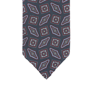 A close-up of a Stefano Cau necktie crafted from luxurious Italian silk, featuring a geometric pattern with red, blue, and white diamond and square shapes on a dark gray background.