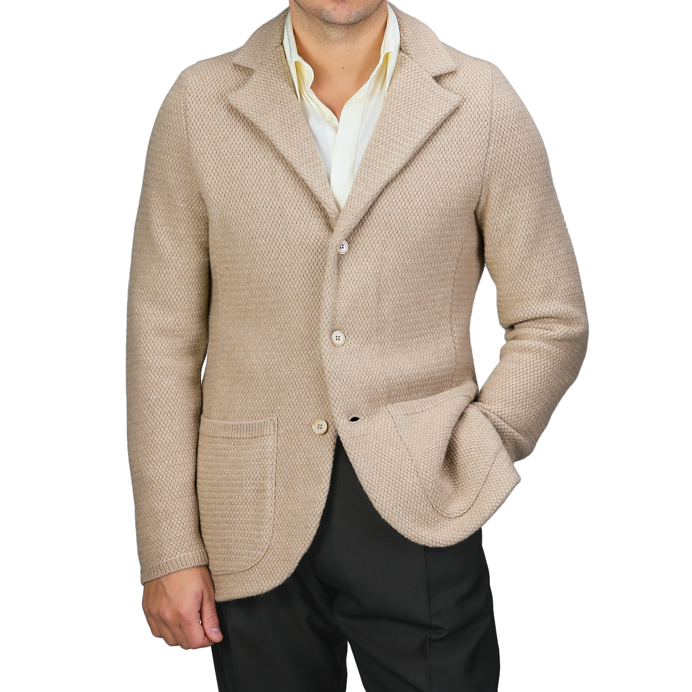 A person wearing a Stenströms Beige Merino Wool Knitted Blazer featuring a tailored waist, paired with a light yellow shirt and dark trousers, poses with one hand in their pocket against a plain backdrop.