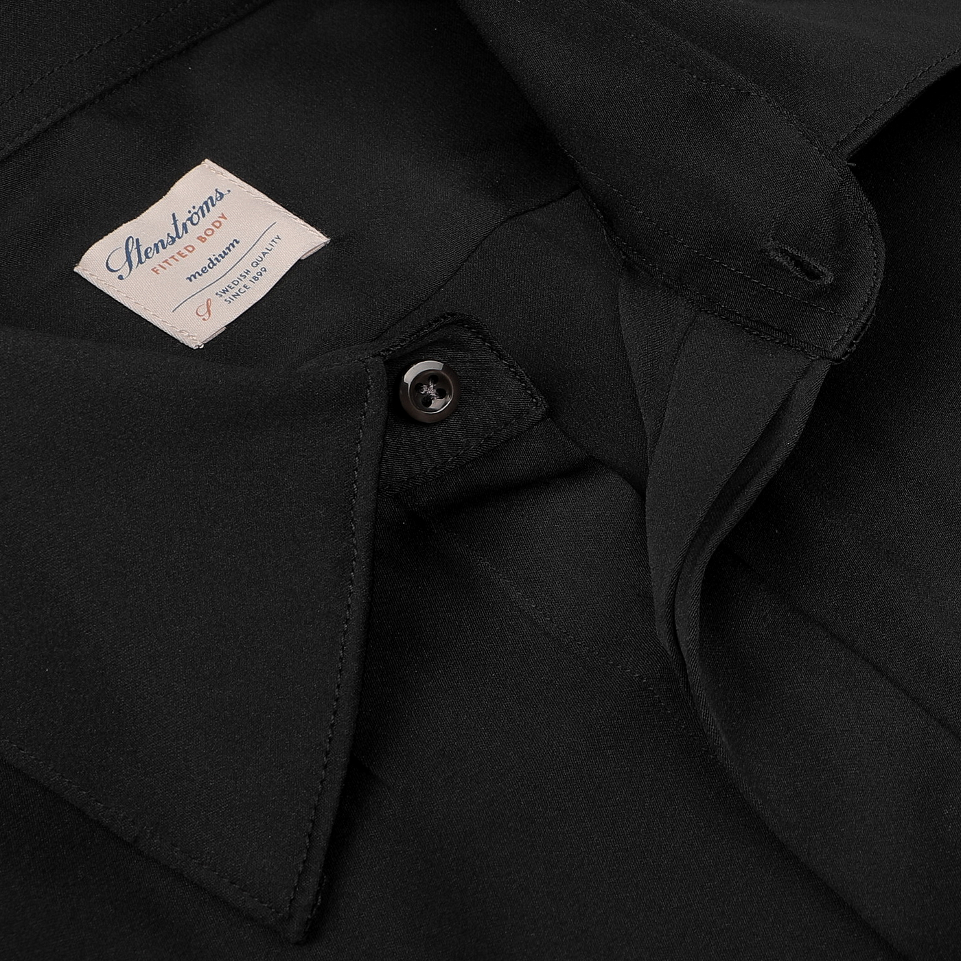 Close-up of the Black Silk Fitted Body Shirt by Stenströms, part of their capsule collection, showcasing a prominently displayed label. This refined piece features a buttoned collar and is expertly folded.