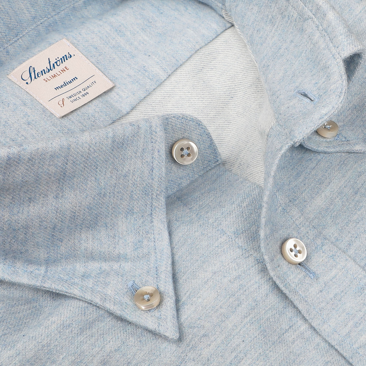 Close-up of a light blue melange flannel Stenströms Light Blue Cotton Flannel Slimline BD Shirt with visible buttons and a tag showing size medium and fabric details. The brushed cotton twill fabric provides a soft texture, and the Slimline cut ensures a sleek fit.