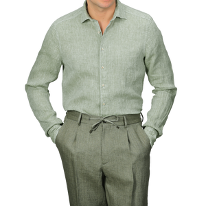 Clad in Stenströms' Light Green Linen Cutaway Slimline Shirt with mother of pearl buttons and coordinated slimline green trousers, a person stands with hands in pockets against a simple background, viewed from the shoulders down.