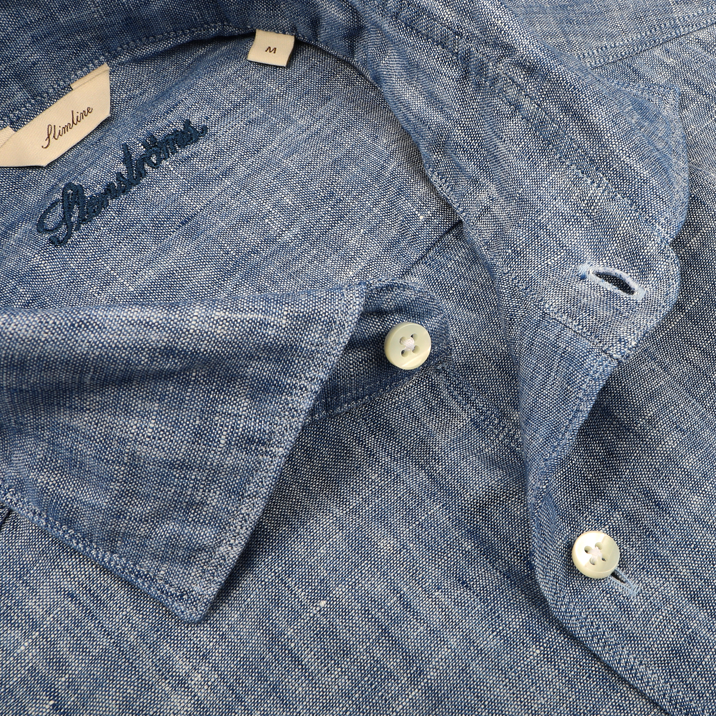 Close-up of a Stenströms Mid Blue Linen Cutaway Slimline Shirt with a textured pattern, collar, and white buttons. The brand label peeks out, making it an ideal summer essential that seamlessly combines style and comfort.