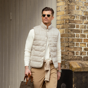 Stylish man in sunglasses wearing a Stenströms beige herringbone cotton linen down padded gilet, sweater, and pants, holding a brown leather bag, standing by a brick wall.