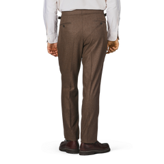 A person wearing high-waist Studio 73 Brown Melange Wool Flannel Pleated Trousers, a white long-sleeve shirt, and dark brown shoes stands with their back to the camera.