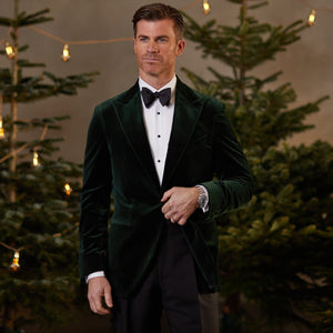 A man in a Dark Green Cotton Velvet Dinner Jacket, crafted by Studio 73 from cotton velvet fabric, standing in front of Christmas trees.