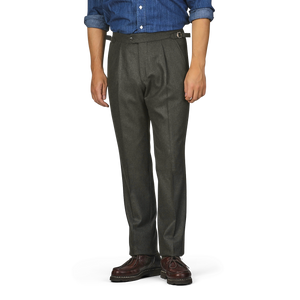 A person is standing wearing Studio 73's dark green wool flannel pleated trousers with a belt, a blue shirt with rolled-up sleeves, and brown shoes. The person's head and lower legs are outside the frame.