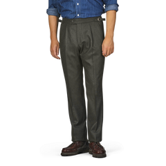 A person is standing wearing Studio 73's dark green wool flannel pleated trousers with a belt, a blue shirt with rolled-up sleeves, and brown shoes. The person's head and lower legs are outside the frame.