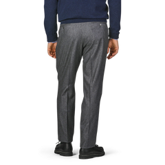 A person wearing Studio 73's Dark Grey Wool Flannel Pleated Trousers and a navy blue sweater is standing with their back facing the camera, showcasing the high-waist tapered leg and overall fit of the trousers.