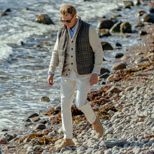 A man wearing Studio 73 Off-White Wool Flannel Pleated Trousers, walks on a rocky beach.