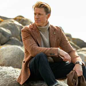 A man, dressed in an Orange Herringbone Wool Tweed Blazer by Studio 73, is sitting on a rock.