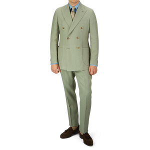 Man wearing a Sage Green Canapa Hemp DB Suit by Studio 73 with a blue shirt, beige tie, and brown shoes, standing against a plain background.