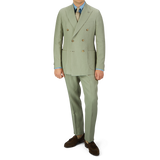 Man wearing a Sage Green Canapa Hemp DB Suit by Studio 73 with a blue shirt, beige tie, and brown shoes, standing against a plain background.