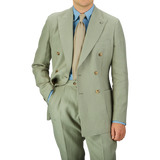 A person wearing a Sage Green Canapa Hemp DB Suit by Studio 73 with a light blue shirt and a beige tie stands with one hand in the pocket.