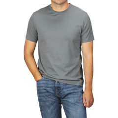 A man is standing against a plain background, wearing a Sunspel Smoke Green Classic Cotton T-Shirt and blue jeans. His left hand is in his jeans pocket. His head is not visible in the photo.