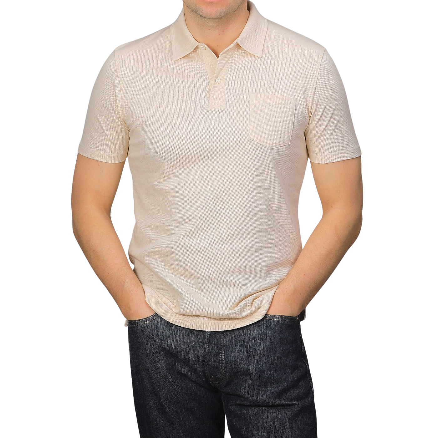 A person in a Sunspel Ecru Beige Cotton Riviera Polo Shirt and dark jeans stands with hands in pockets against a plain grey background.