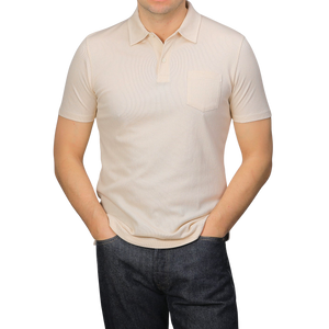 A person in a Sunspel Ecru Beige Cotton Riviera Polo Shirt and dark jeans stands with hands in pockets against a plain grey background.