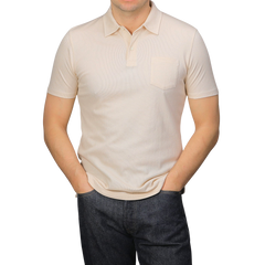 A person in a Sunspel Ecru Beige Cotton Riviera Polo Shirt and dark jeans stands with hands in pockets against a plain grey background.