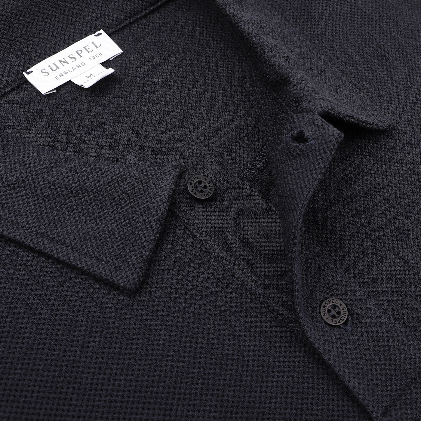 Close-up of a Midnight Blue Riviera Polo Shirt by Sunspel, crafted from premium cotton mesh fabric. The label reads "Sunspel, England 1860," reflecting the timeless style favored by icons like Daniel Craig.