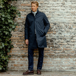 A man in a Sunspel Navy Blue Cotton Mac Car Coat leaning against a brick wall.
