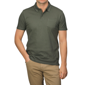 Dressed in a Sunspel Pine Green Cotton Riviera Polo Shirt with a chest pocket, paired with beige pants, one casually slips a hand in a pocket, epitomizing the contemporary and sophisticated style of its breathable cotton fabric.