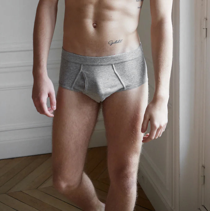 A person stands indoors, wearing **The White Briefs Grey Melange Pima Cotton Platan Briefs**. The lower body and part of the torso are visible, with a tattoo on the right side of the abdomen.