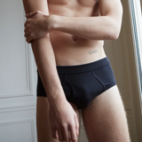 A shirtless person adjusts their arm, wearing The White Briefs Parisian Night Pima Cotton Platan Briefs, standing indoors near a white paneled wall and a window on a Parisian night.