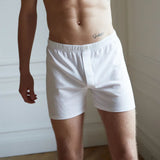 A person wearing The White Briefs White Lyocell Cotton Henner Boxers stands in a room with white walls and a wooden floor.