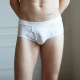 A person wearing White Pima Cotton Platan Briefs from The White Briefs stands against a white wall with hands behind their back.