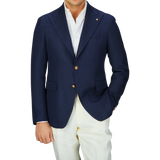 A person wearing a Navy Blue Herringbone Cotton Linen Vesuvio Blazer by Tagliatore with peak lapel and white pants against a gray background.