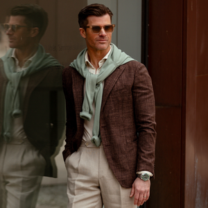A stylish man in a Tagliatore Dark Brown Melange Wool Linen Silk Blazer and sweater leans against a glass window, his reflection visible.