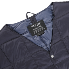 Close-up of a navy blue quilted jacket with a visible clothing tag labeled "Taion." This nylon down padded jacket, crafted from wind-resistant fabric, features a snap button closure and a hanging loop.