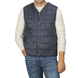 A man is wearing the Taion Navy Blue Nylon Down Padded Vest over a white sweater and blue jeans, standing against a plain background. The vest’s high-density nylon material adds an element of sophistication to his look.